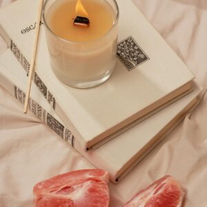 High angle of burning aroma candle with incense stick on books near pieces of ripe grapefruit on creased fabric