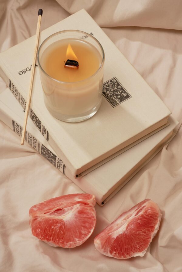 High angle of burning aroma candle with incense stick on books near pieces of ripe grapefruit on creased fabric